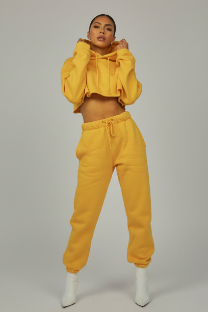 A model stands in a studio wearing a matching bright yellow cropped hoodie and jogger sweatpants with white ankle boots.